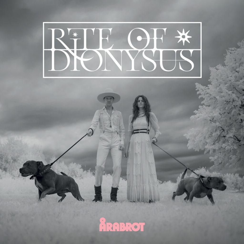 Albumcover Årabrot – Rite Of Dionysus: Schwarz-weiß Fotografie, Kjetil Nernes im Cowboy-Outfit mit Hut und Karin Park im weißen Kleid führen zwei bullige Hunde an Leinen. Surreale, kontrastreiche Landschaft mit wolkigem Himmel, darüber Albumtitel in geometrischer Typografie.