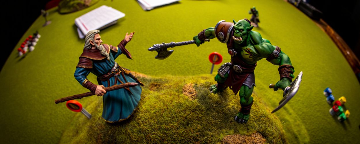 Miniaturen von einem orkischen Krieger und einem Elfen-Zauberer kämpfen auf einem Spieltisch mit Würfeln, Karten und Gelände – Warcrow Starter-Set.