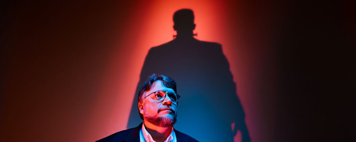 Guillermo Del Toro steht im blauen und roten Licht eines Kinosaals. Hinter ihm erscheint die überlebensgroße Silhouette von Frankensteins Kreatur – Sinnbild seines Kampfes gegen künstliche Intelligenz.