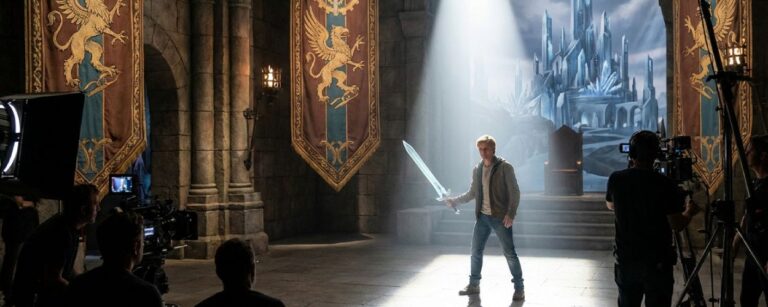 Auf einem Filmset in einer hallenartigen Thronsaal-Kulisse mit großen Bannern und Fackeln steht ein junger Mann in Jeans und Hoodie im Lichtkegel und hält ein leuchtendes Schwert, umgeben von Kameraleuten, im Hintergrund ist eine gemalte Kristallburg zu sehen – Symbolbild für das moderne He-Man-Reboot Masters of the Universe.