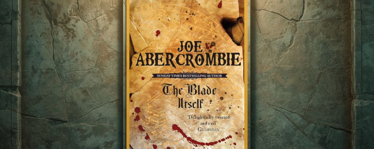 Vor einer rissigen, moosig wirkenden Steinwand hängt das Cover von Joe Abercrombies The Blade Itself in einem goldenen Rahmen. Die Gestaltung zeigt Pergamentoptik, feine Linien wie eine Karte und rote Blutspritzer, wodurch das Motiv wie ein ausgestelltes Relikt wirkt.