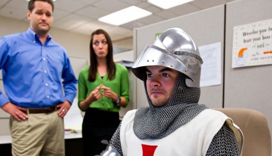 Ein Kreuzritter in Rüstung sitzt im Büro am Computer; im Hintergrund stehen zwei Kollegen. Unten steht der Text über „Ethos“ und Lieferdienst.