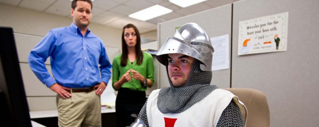 Ein Kreuzritter in Rüstung sitzt im Büro am Computer; im Hintergrund stehen zwei Kollegen. Unten steht der Text über „Ethos“ und Lieferdienst.