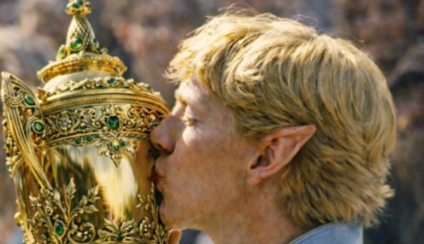 Analoge Fantasy-Sportfotografie im Hochformat: Ein blonder Hochelf mit spitzen Ohren und himmelblauer, goldbestickter Zeremonien-Sportkleidung küsst einen prunkvollen goldenen Pokal mit grünen Edelsteinen. Im Hintergrund verschwimmt die Menge wie bei einem historischen Turniermoment.