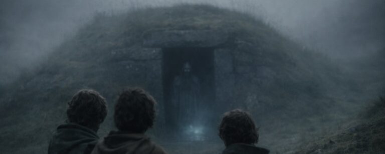 Drei Hobbits stehen im Nebel vor alten Hügelgräbern, während hinter ihnen ein geisterhafter Schatten aus einem Grabgang zu kriechen scheint.
