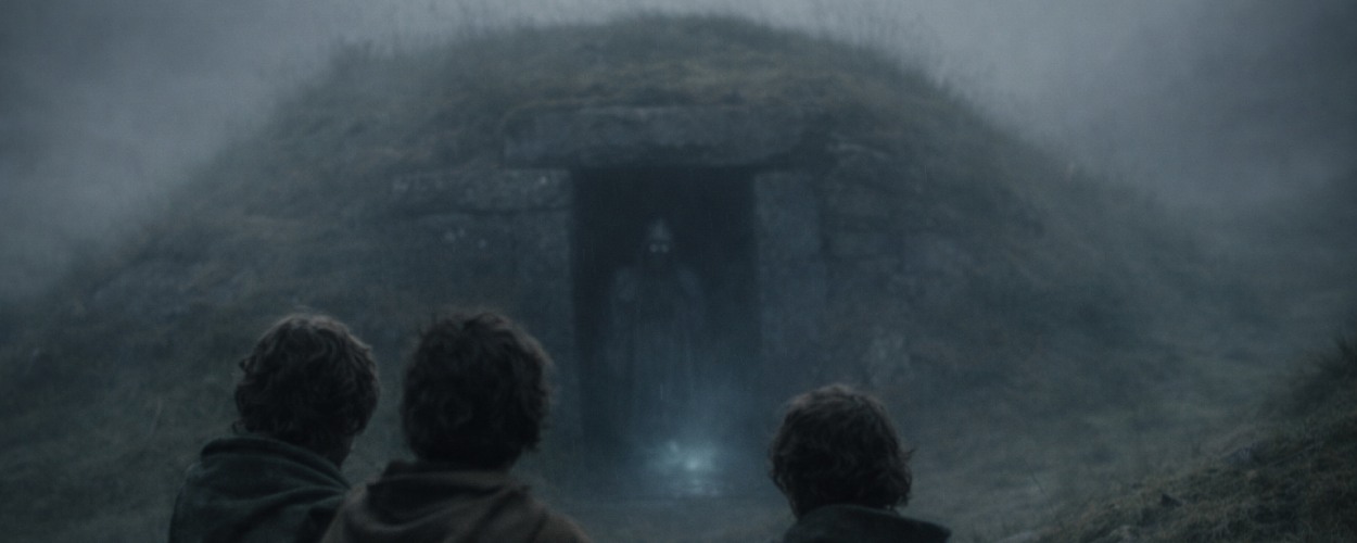 Drei Hobbits stehen im Nebel vor alten Hügelgräbern, während hinter ihnen ein geisterhafter Schatten aus einem Grabgang zu kriechen scheint.