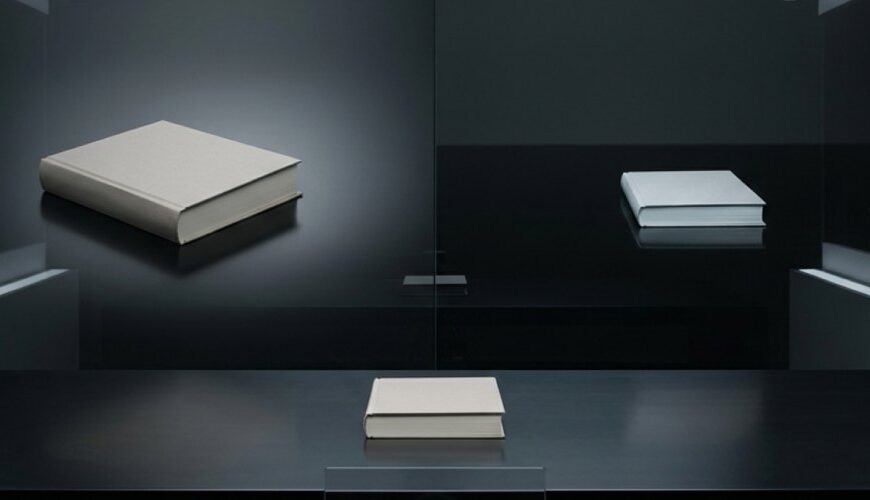Ein einzelnes Buch liegt in einem kalten Studioraum vor einer Kamera, während es im dunklen Hintergrund zweimal unterschiedlich erscheint: einmal größer und feierlicher, einmal kleiner und sichtbar herabgesetzt.