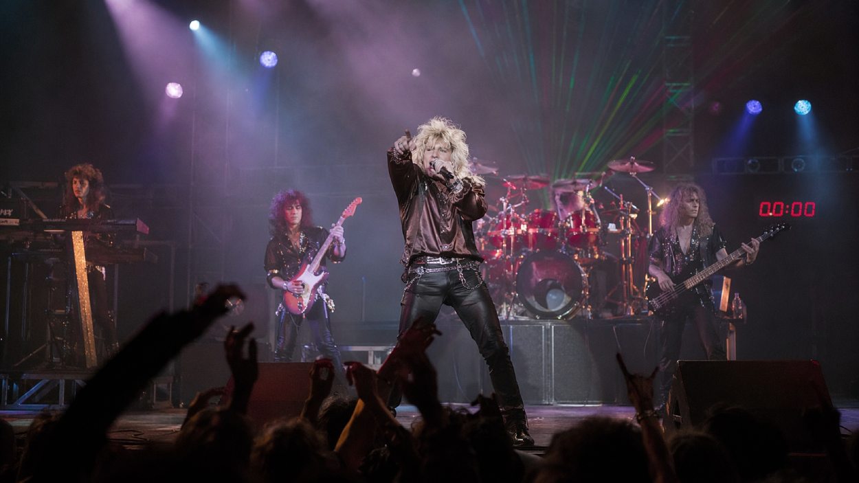 80er-Rockband auf einer Konzertbühne mit blondem Sänger in heroischer Pose, Synthesizer, Schlagzeug, Lasern und rotem Countdown-Display im Hintergrund.