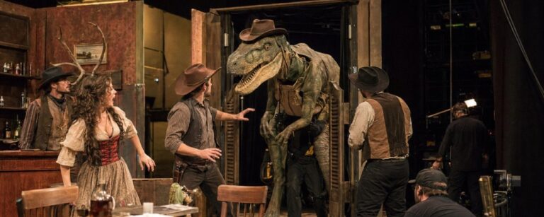 Dokumentarisch wirkende Theaterprobe auf einer Western-Saloon-Bühne: Ein großer Dinosaurier mit Cowboyhut tritt durch Schwingtüren, während Schauspieler in Westernkostümen und Bühnentechniker die absurde Szene beobachten.