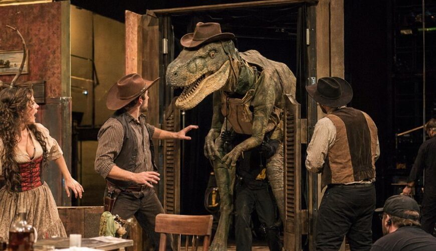 Dokumentarisch wirkende Theaterprobe auf einer Western-Saloon-Bühne: Ein großer Dinosaurier mit Cowboyhut tritt durch Schwingtüren, während Schauspieler in Westernkostümen und Bühnentechniker die absurde Szene beobachten.