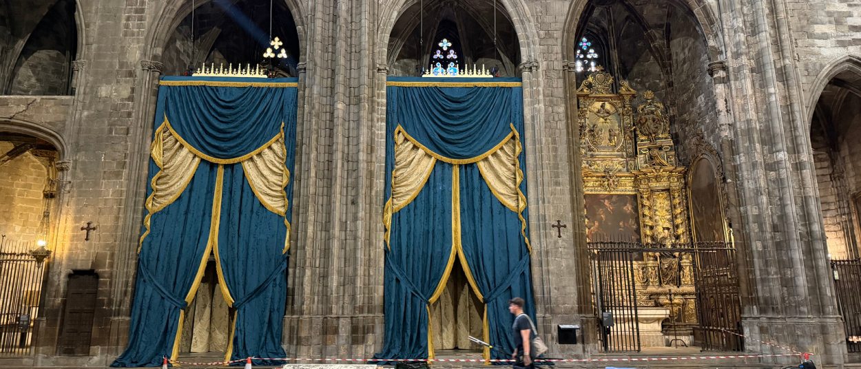 Zwei hohe blau-goldene Vorhänge hängen zwischen den steinernen Bögen der Kathedrale von Girona, während ein Mann an Absperrbändern vorbeigeht.