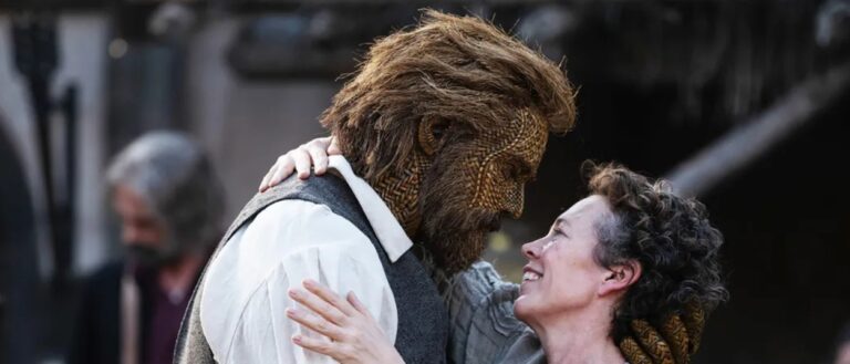 Olivia Colman umfasst auf einem Dorfplatz einen großen Mann mit einem vollständig aus geflochtener Weide gefertigten Gesicht.