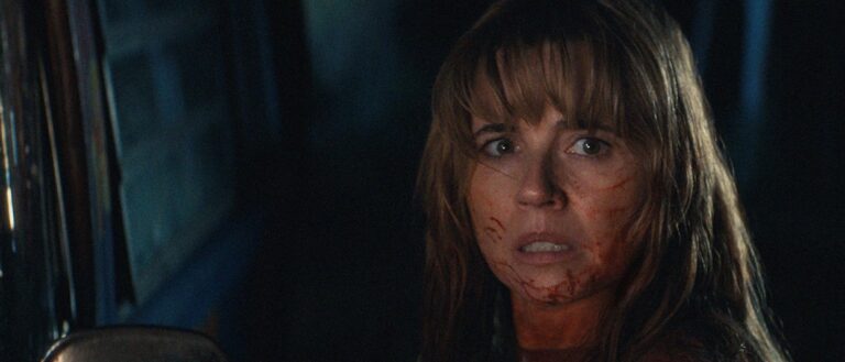 Pamela Voorhees steht mit ernstem Blick vor einem bewaldeten See, während dunkle Äste und die Wasseroberfläche die düstere Szene einrahmen.