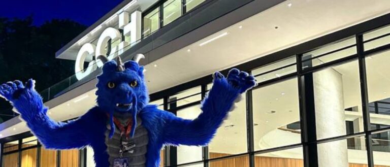 Ein Fursuiter in einem großen blauen Tierkostüm hebt vor dem beleuchteten Eingang des Congress Center Hamburg beide Arme in die Höhe.