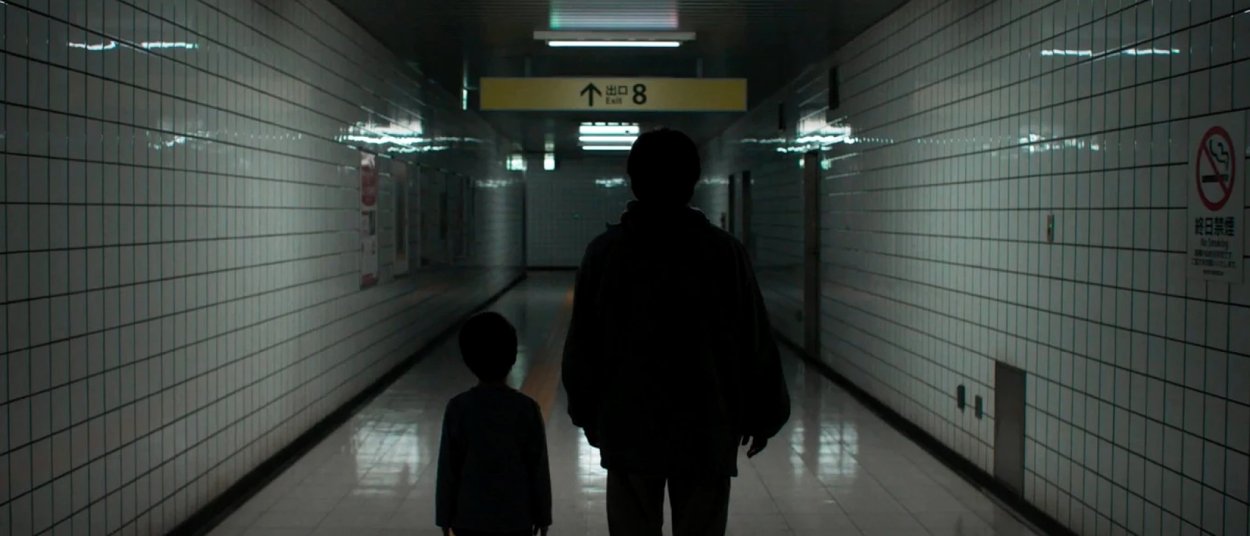 Ein erwachsener Mann und ein Kind gehen durch einen dunklen gefliesten U-Bahn-Gang auf ein gelbes Schild mit der Nummer 8 zu.