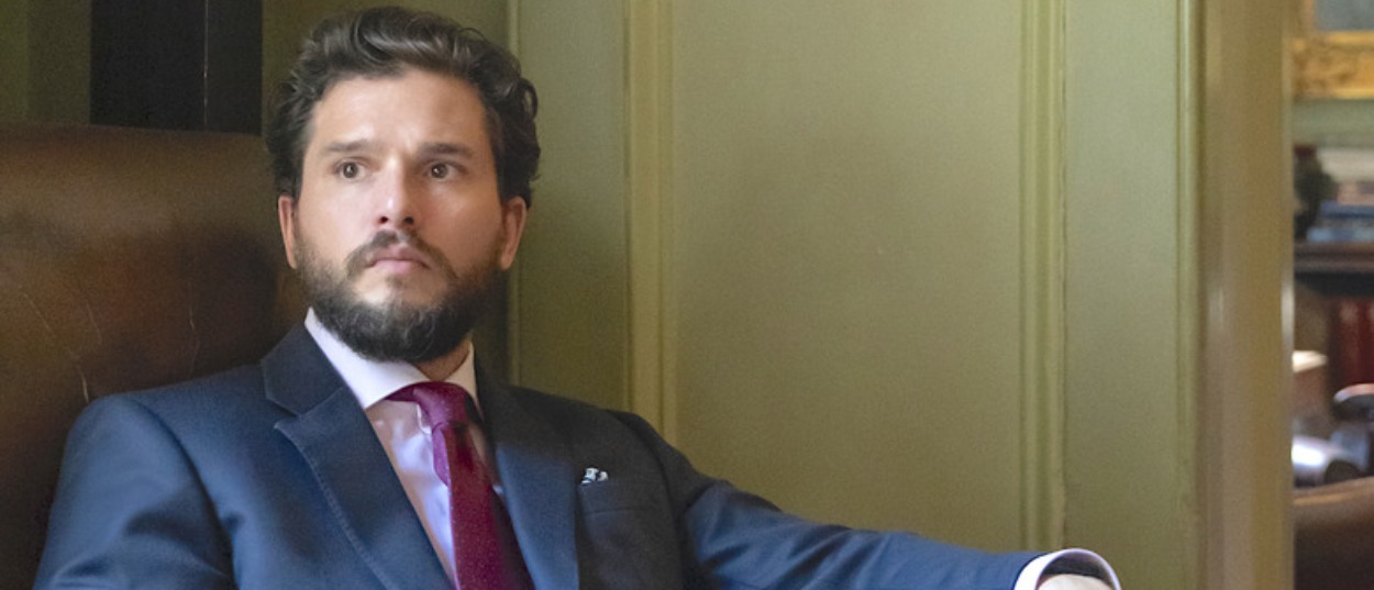 Kit Harington sitzt im dunklen Anzug und mit roter Krawatte in einem Ledersessel und blickt zur Seite.