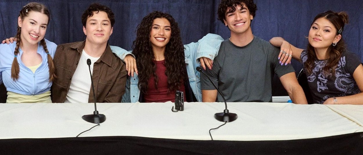 Eva Smedley, Harry Jones, Dior Goodjohn, Jacob Rodriguez und Momona Tamada sitzen nebeneinander hinter einem Tisch und lächeln in die Kamera.