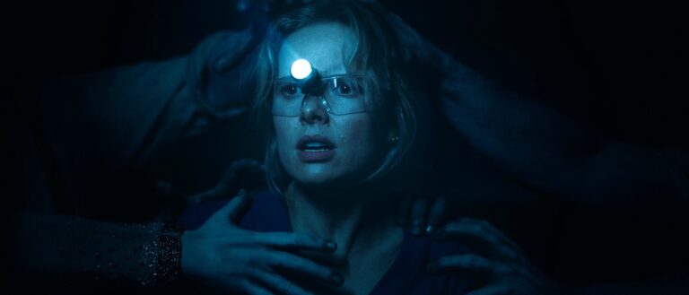 Gemma trägt eine Stirnlampe und Schutzbrille und blickt erschrocken in die Dunkelheit. Mehrere blasse Hände greifen von hinten und von den Seiten nach ihrem Kopf, Hals und ihren Schultern.