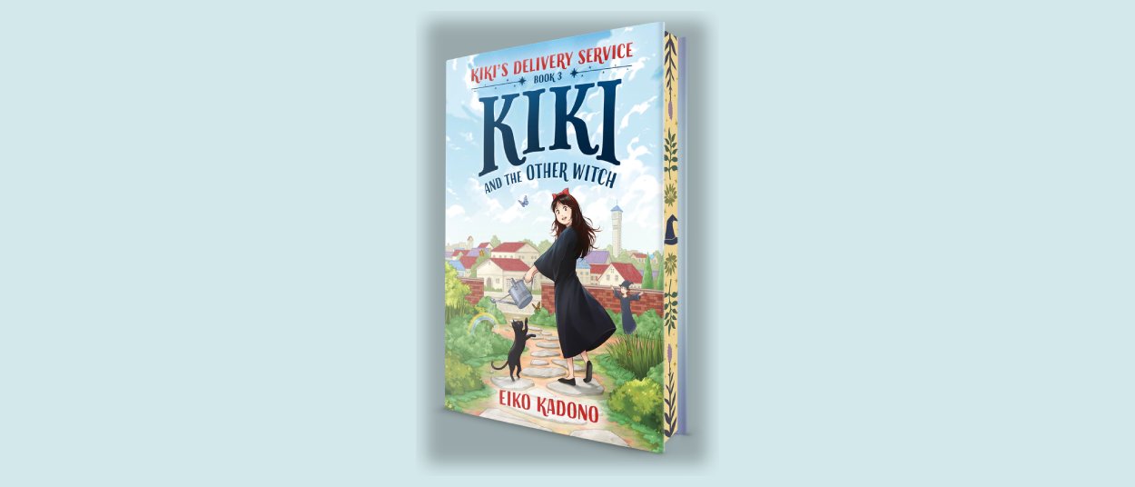 Das Buchcover von Kiki and the Other Witch zeigt Kiki mit schwarzem Kleid und roter Schleife neben dem schwarzen Kater Jiji auf einer grünen Anhöhe über einer hellen Küstenstadt.
