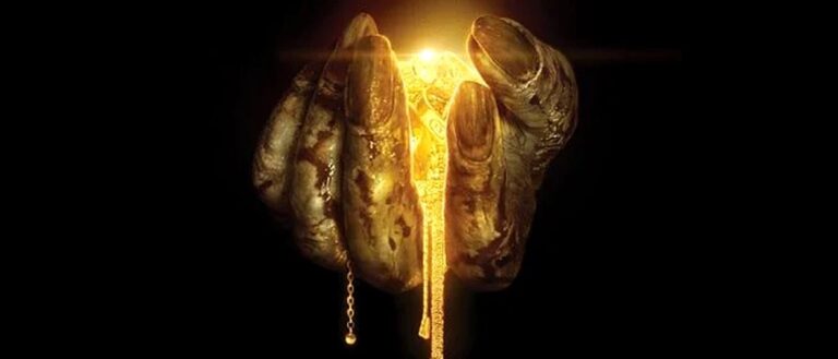 Eine schmutzige, krallenartige Hand hält mehrere goldene Schmuckstücke vor schwarzem Hintergrund. Zwischen den Fingern leuchtet helles goldenes Licht.