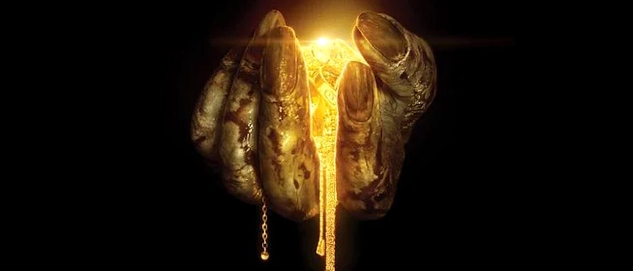 Eine schmutzige, krallenartige Hand hält mehrere goldene Schmuckstücke vor schwarzem Hintergrund. Zwischen den Fingern leuchtet helles goldenes Licht.