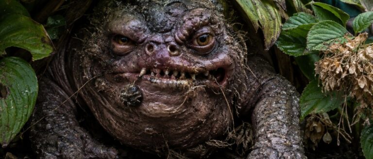 Makroartiges Fantasy-Naturfoto im Hochformat: Ein feister, krötenartiger Beethocker mit schlammiger Haut, schiefen Zähnen, gelben Augen und langen Krallen lauert unter breiten Blättern in einem gepflegten Vorgarten. Neben ihm stehen ein Gartenzwerg, helle Ziersteine und nasse Pflanzen. Das Wesen wirkt wie ein erschreckend echter, bösartiger Gartenfund aus den Zwischenreichen.