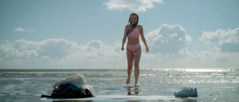 Eine junge Frau in pinkfarbener Badebekleidung steht im flachen Meerwasser unter blauem Himmel; im Vordergrund liegen mehrere Gegenstände im Wasser.