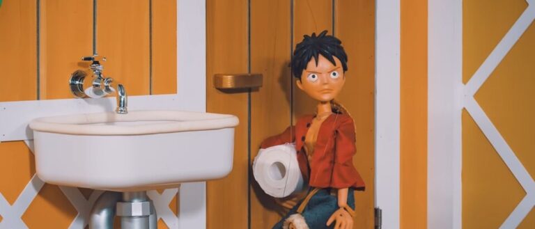 Die Ruffy-Marionette steht mit einer großen Rolle Toilettenpapier neben einem weißen Waschbecken in einem orange gestrichenen Raum der Thousand Sunny.