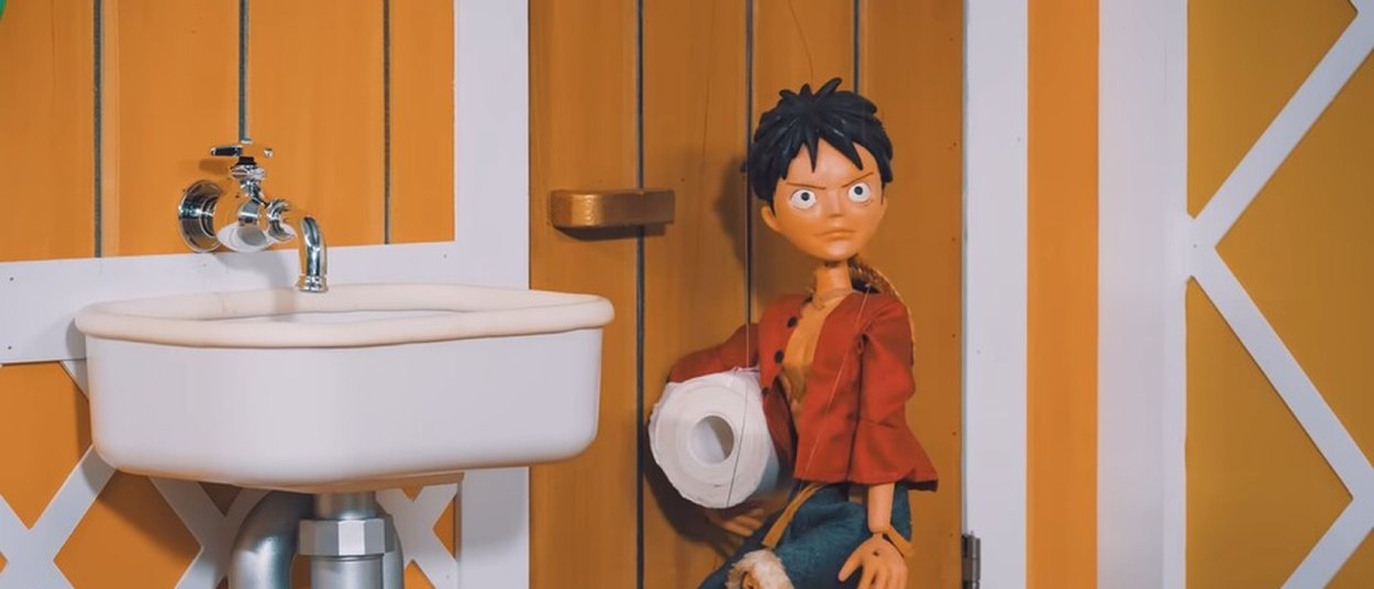 Die Ruffy-Marionette steht mit einer großen Rolle Toilettenpapier neben einem weißen Waschbecken in einem orange gestrichenen Raum der Thousand Sunny.