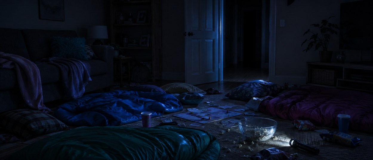 Mehrere Schlafsäcke liegen nachts in einem dunklen Wohnzimmer zwischen Snackresten, einem Brettspiel und einer Taschenlampe, während eine halb offene Tür in einen schwarzen Flur führt.