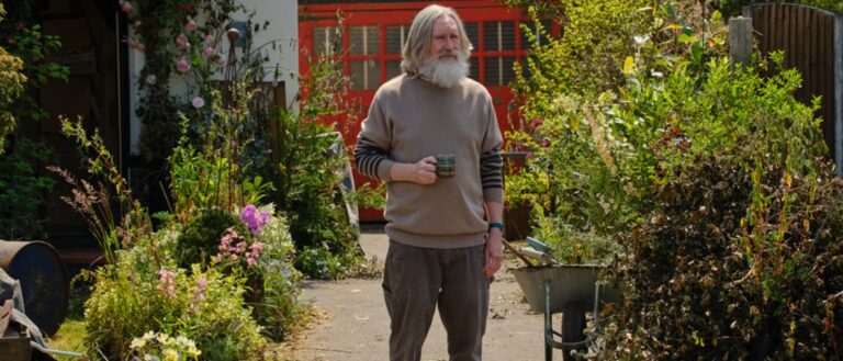 Ein älterer grauhaariger Mann mit Bart steht mit einer Tasse in einem dicht bewachsenen britischen Garten zwischen Blumen, Schubkarre und einem roten Gartengebäude.