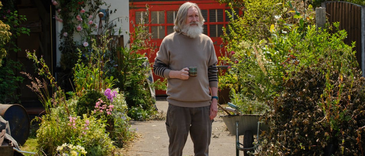 Ein älterer grauhaariger Mann mit Bart steht mit einer Tasse in einem dicht bewachsenen britischen Garten zwischen Blumen, Schubkarre und einem roten Gartengebäude.