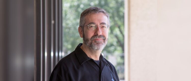 Warren Spector steht mit grauem Bart und Brille in einem schwarzen Hemd vor einer großen Fensterfront und blickt lächelnd in die Kamera.