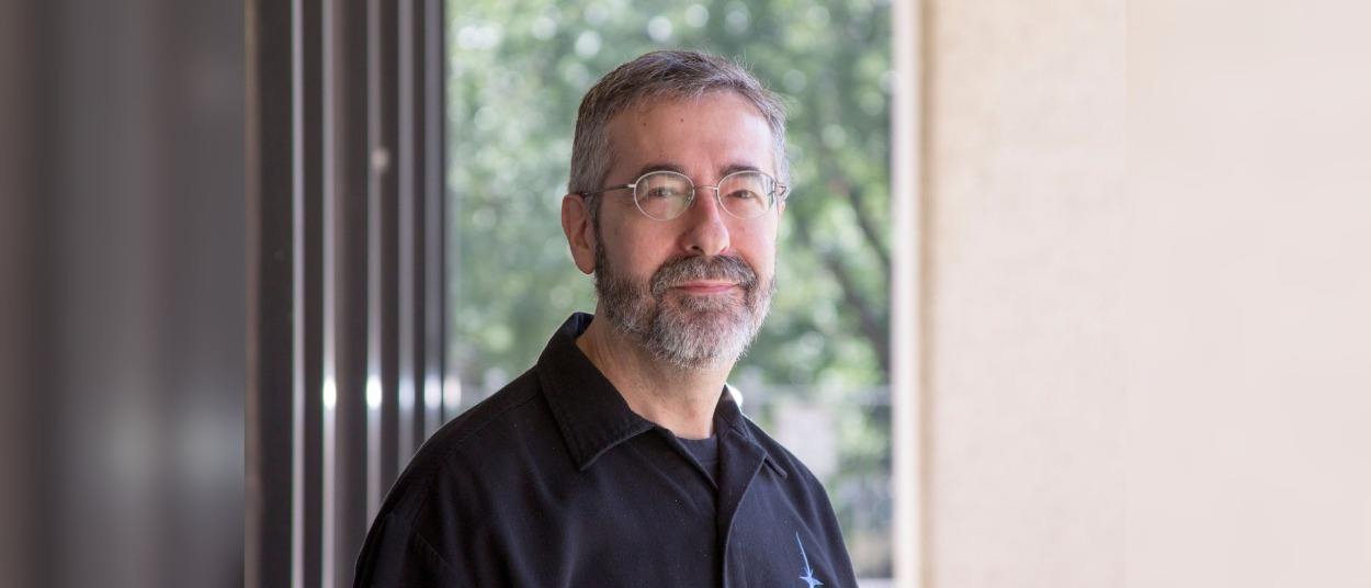Warren Spector steht mit grauem Bart und Brille in einem schwarzen Hemd vor einer großen Fensterfront und blickt lächelnd in die Kamera.