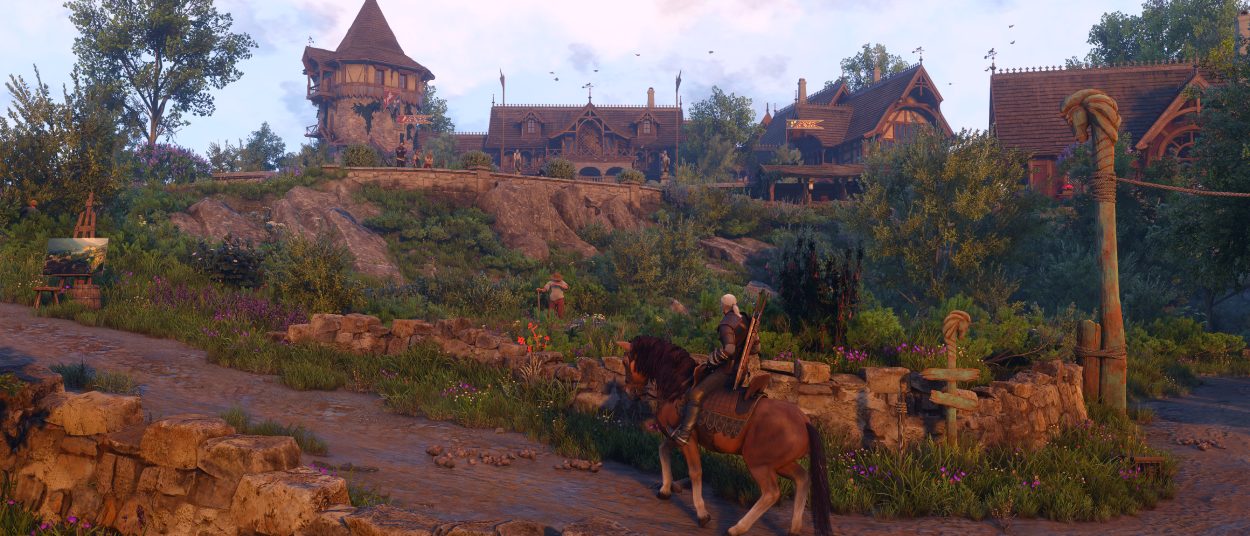 Ein weißhaariger Hexer reitet auf einem Pferd durch eine üppige Landschaft auf eine große mittelalterliche Siedlung mit Fachwerkhäusern, Turm und blühenden Wiesen zu.
