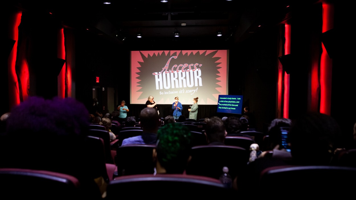 Publikum in einem Kinosaal beim Access:Horror Film Festival; auf der Bühne sprechen mehrere Personen, daneben läuft eine separate Untertitelanzeige.