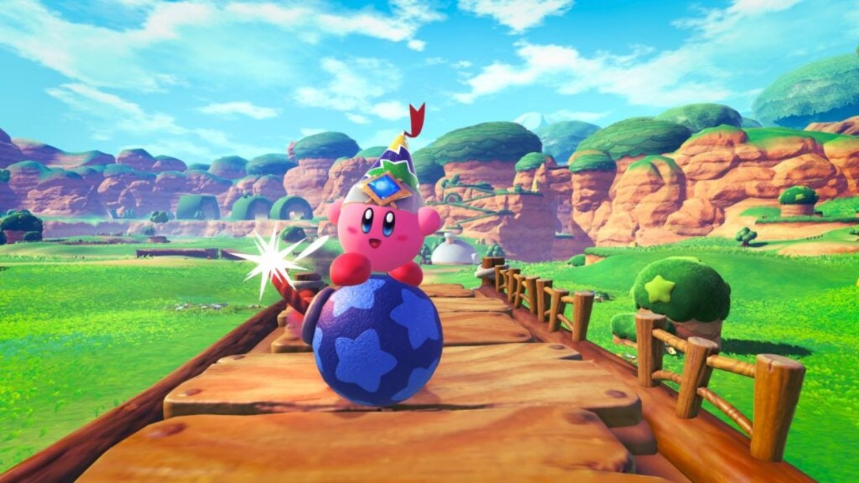 Kirby sitzt in Kirby and the World Beyond auf einer großen blauen Kugel und rollt über einen Holzweg durch eine farbenfrohe offene Landschaft.