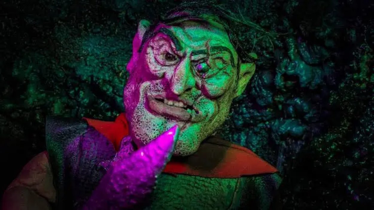 Eine groteske handgefertigte Puppenfigur aus Kylgor in the Grisly Abyss wird in einer dunklen Höhle von grünem und violettem Licht angestrahlt.
