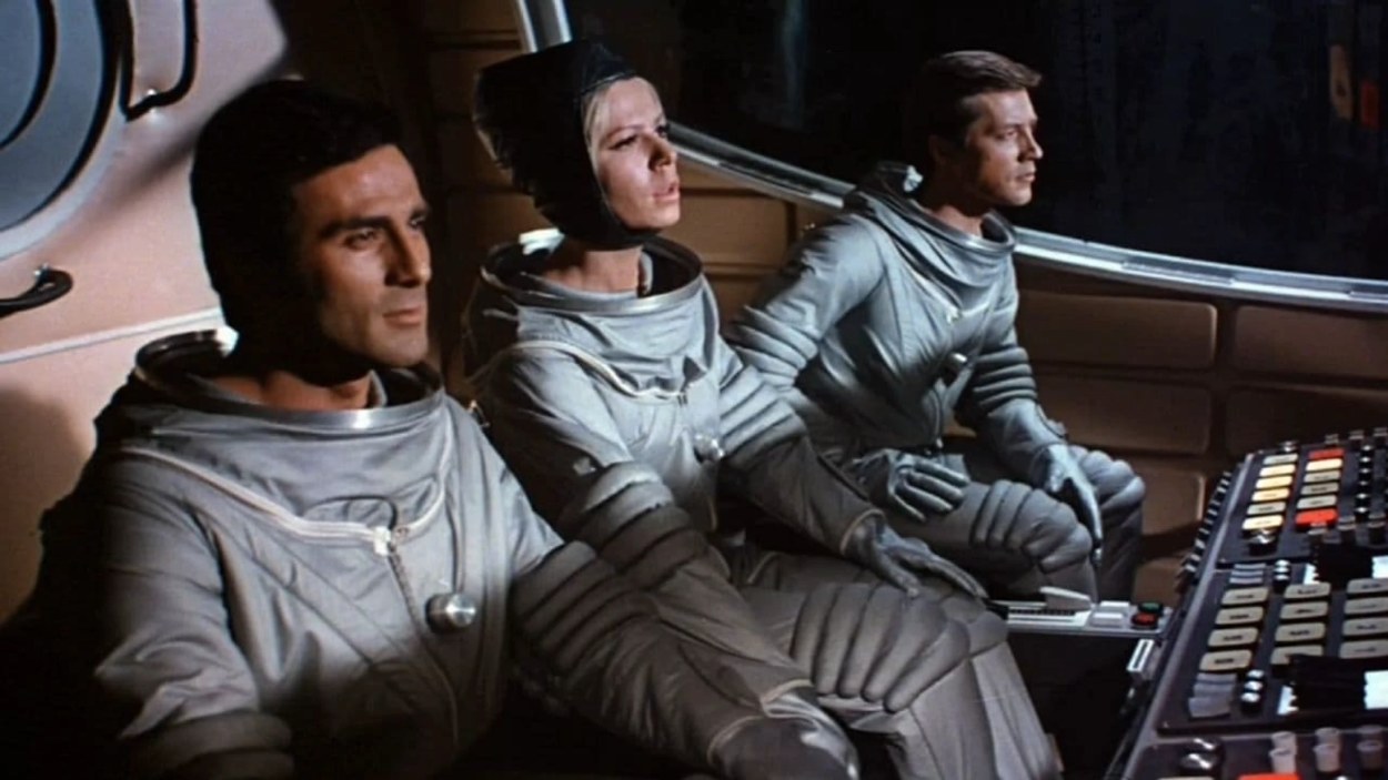 Drei Besatzungsmitglieder aus Signale – Ein Weltraumabenteuer sitzen in grauen Raumanzügen im Cockpit eines futuristischen Raumschiffs vor einer Konsole.