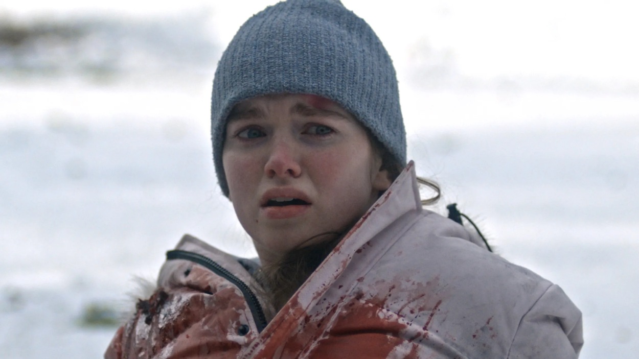 Verängstigte junge Frau mit grauer Mütze und blutverschmierter Winterjacke in verschneiter Umgebung im Horrorfilm Sundown.