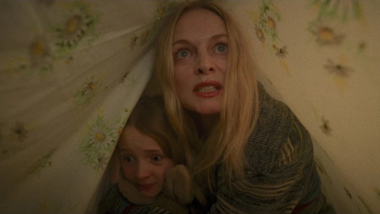 Heather Graham kauert mit einem Kind unter einem geblümten Bettlaken und blickt angespannt nach vorn, während beide Schutz in dem engen Versteck suchen.