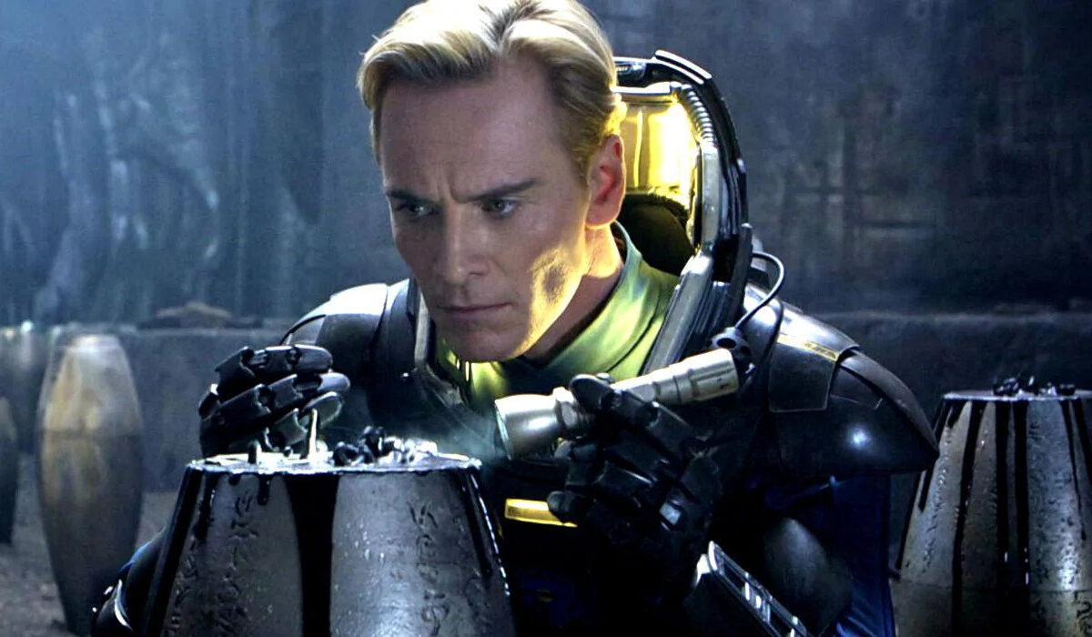 Filmszene aus Prometheus: Ein blondhaariger Mann in einem dunklen futuristischen Schutzanzug untersucht konzentriert einen geheimnisvollen metallischen Behälter. Kaltes blaues Licht und eine düstere außerirdische Umgebung prägen die Szene.