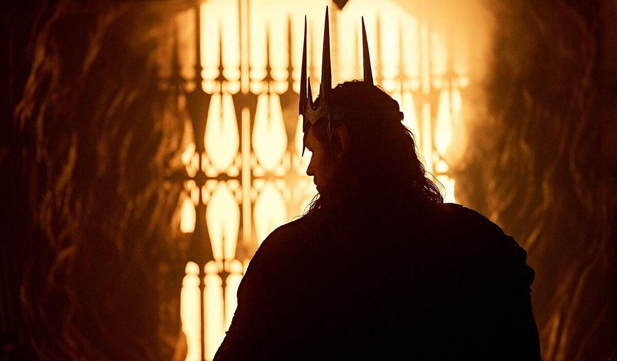 Sauron steht als dunkle Silhouette vor einem grell leuchtenden, feurigen Hintergrund. Er trägt eine spitz aufragende Krone und blickt seitlich in einen glühenden Raum.
