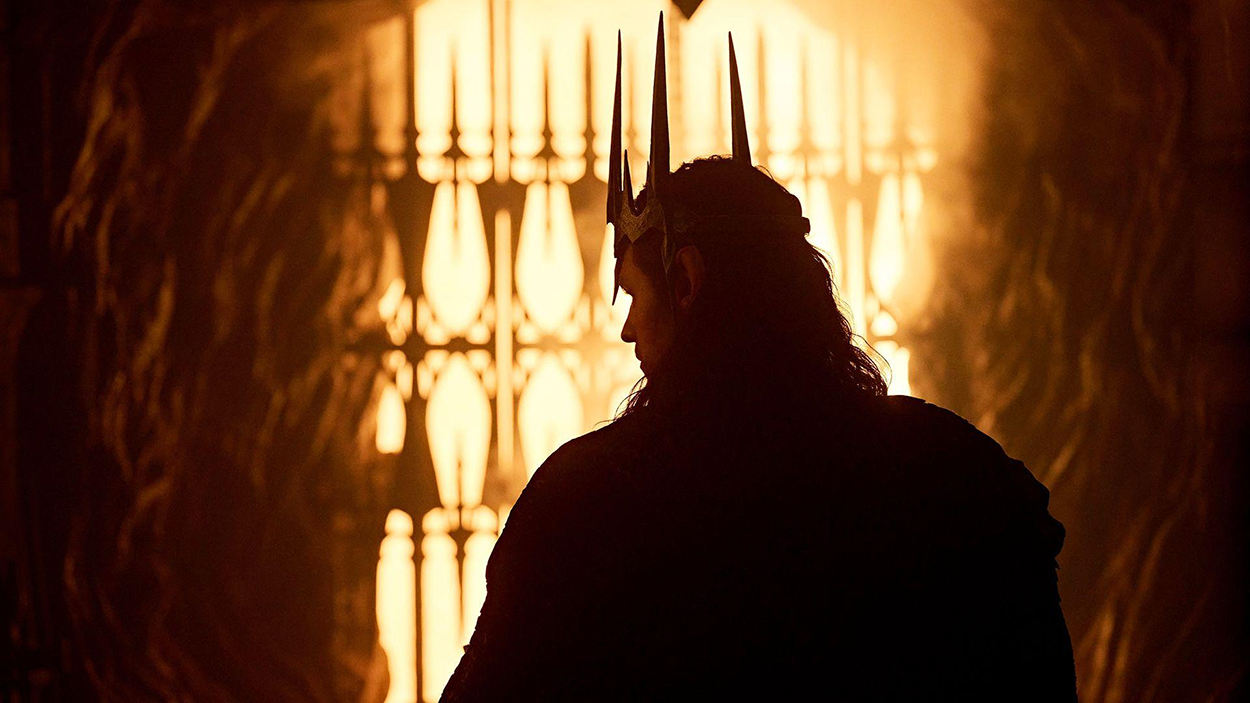 Sauron steht als dunkle Silhouette vor einem grell leuchtenden, feurigen Hintergrund. Er trägt eine spitz aufragende Krone und blickt seitlich in einen glühenden Raum.