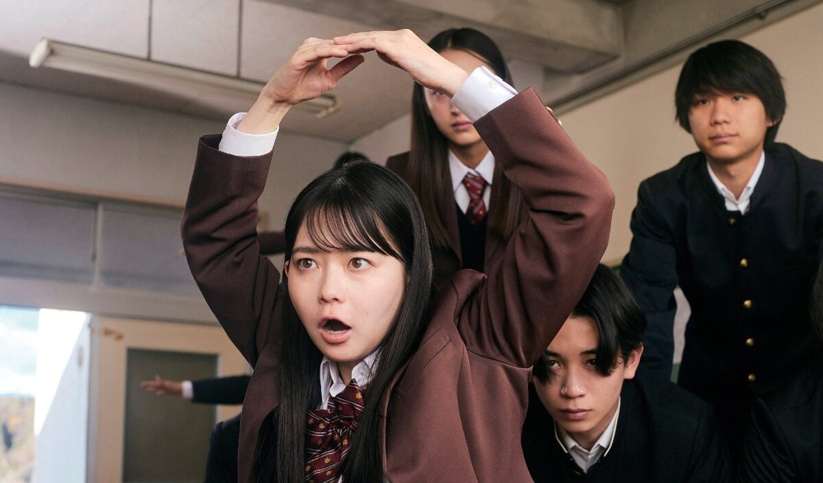 Eine japanische Schülerin hebt in New Group während einer Turnübung die Arme über den Kopf, während mehrere Mitschüler sie aufmerksam beobachten.