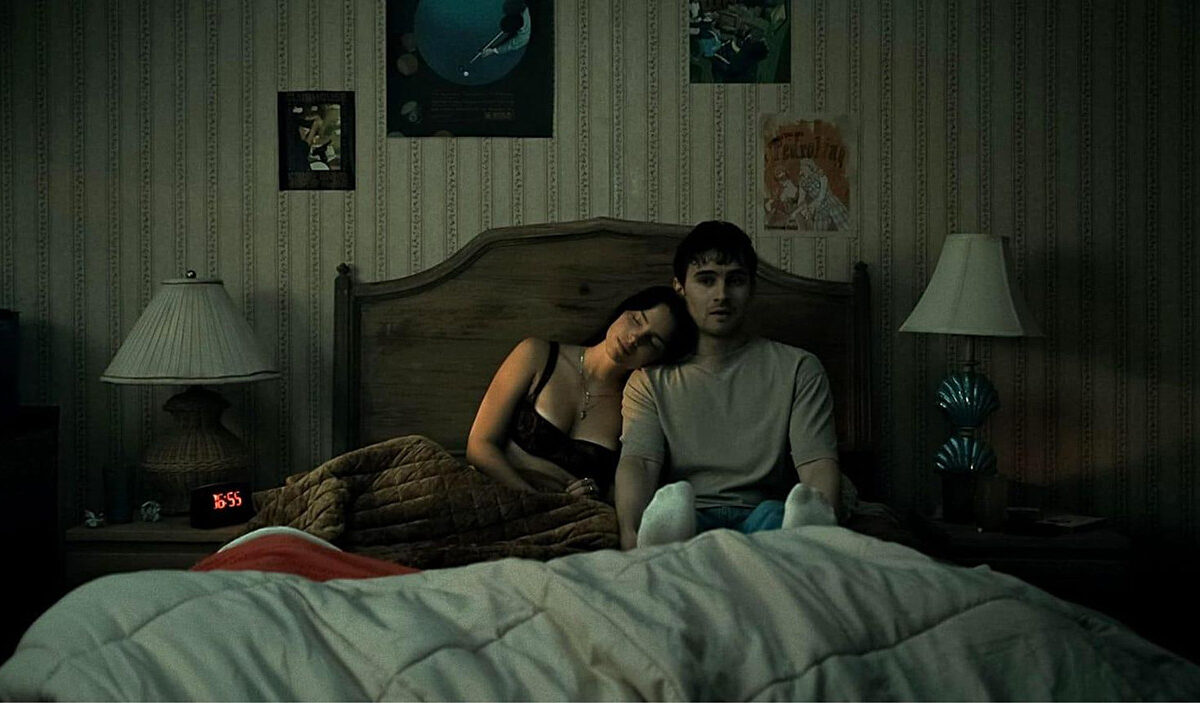 Düstere Schlafzimmerszene aus dem Horrorfilm Obsession von Curry Barker. Eine dunkelhaarige Frau lehnt mit geschlossenen Augen an der Schulter eines jungen Mannes, der mit starrem, ausdruckslosem Blick auf einem Bett sitzt. Gedämpftes grünliches Licht und altmodische Tapeten erzeugen eine beklemmende Atmosphäre.