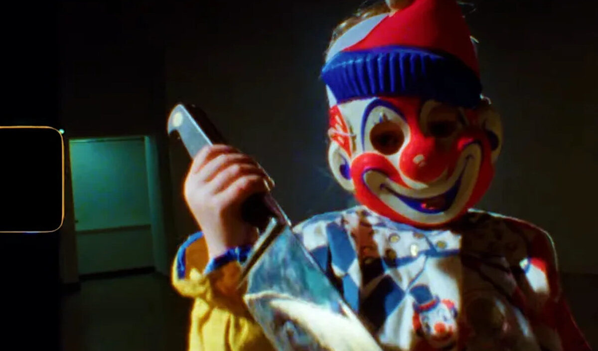 Unheimliche Gestalt mit rot-weiß-blauer Clownsmaske und großem Messer in einem dunklen Raum. Das körnige Filmbild aus Project Shamrock wirkt wie eine beschädigte Archivaufnahme aus den 1960er-Jahren.