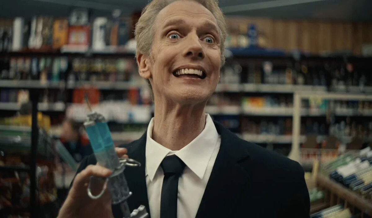 Doug Jones als unheimlicher Grin Man im Horrorkurzfilm Remain Calm. Der Schauspieler steht mit weit aufgerissenen Augen und einem übertrieben breiten Grinsen in einer Tankstelle. Er trägt einen schwarzen Anzug mit Krawatte und hält einen kleinen Gegenstand in der Hand.