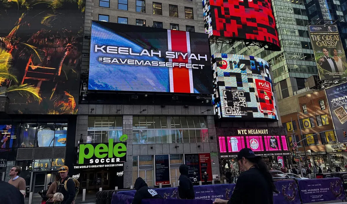Foto vom New Yorker Times Square mit einer großen digitalen Werbetafel, auf der über einem blau-roten Science-Fiction-Motiv die Schriftzüge „KEELAH SI'YAH“ und „#SAVEMASSEFFECT“ zu sehen sind. Darunter befinden sich Geschäfte und Passanten.