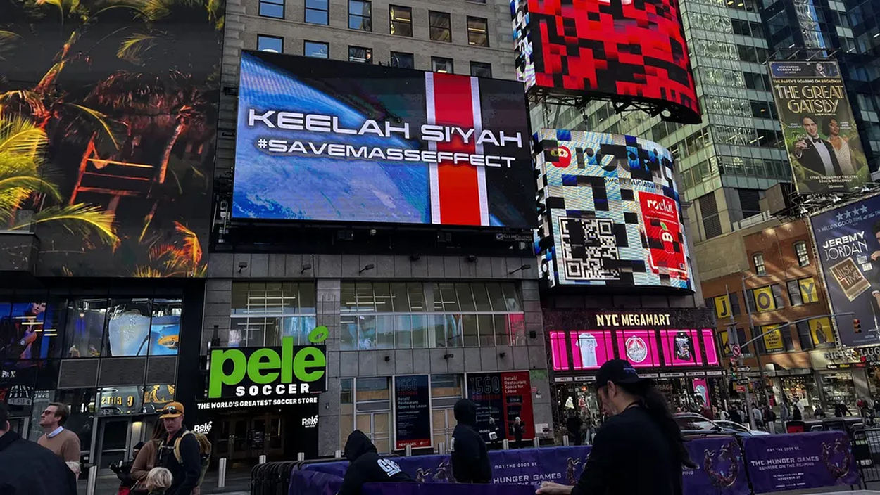 Foto vom New Yorker Times Square mit einer großen digitalen Werbetafel, auf der über einem blau-roten Science-Fiction-Motiv die Schriftzüge „KEELAH SI'YAH“ und „#SAVEMASSEFFECT“ zu sehen sind. Darunter befinden sich Geschäfte und Passanten.